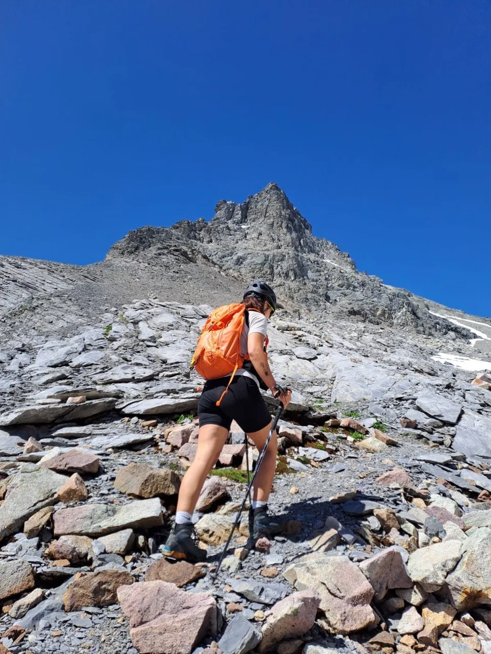 A hiker with an orange backpack crosses a scree field toward a striking rocky summit.