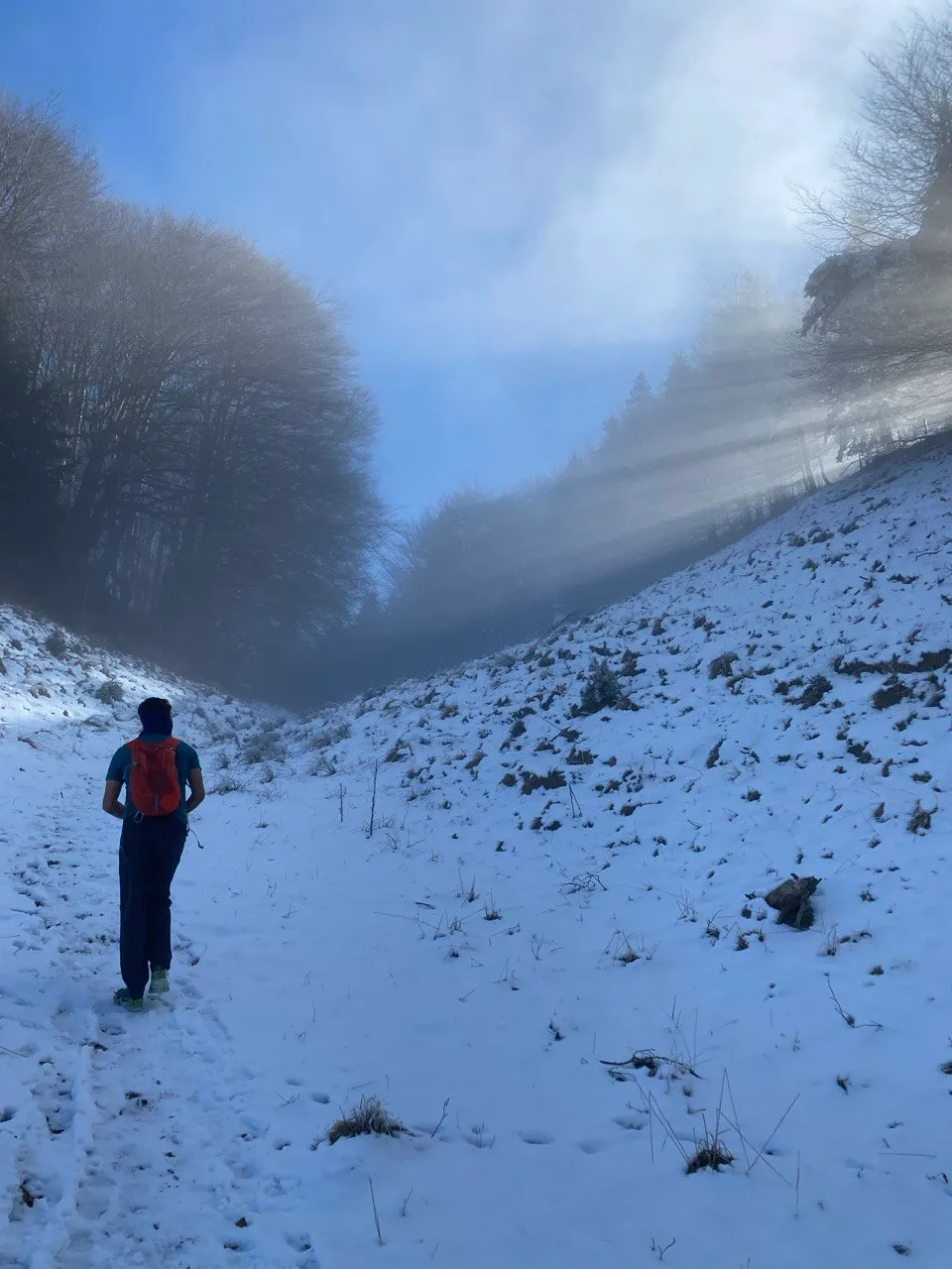 A hiker walks through snow along a ridge as sunbeams break through the fog.