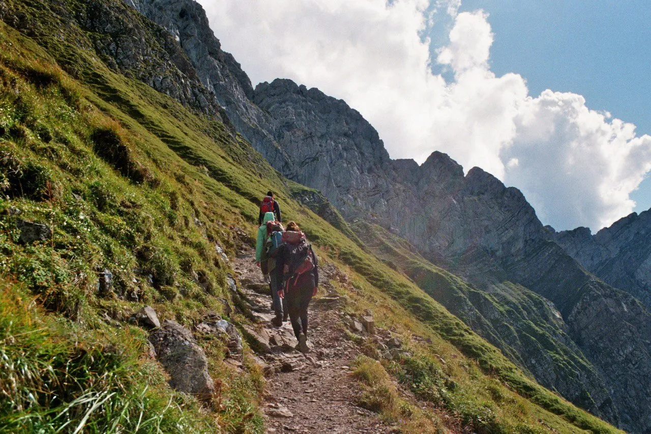 A group of hikers climbing a narrow grassy path toward the rocky summit ridge of Pilatus, cumulus clouds towering alongside.