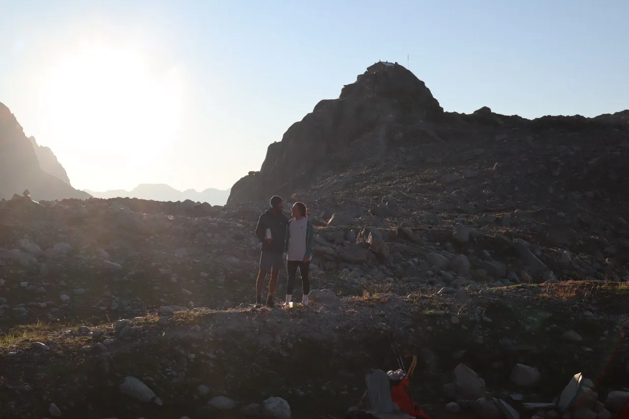 A couple stands among the boulders at sunset, the summit and its hut silhouetted behind them.
