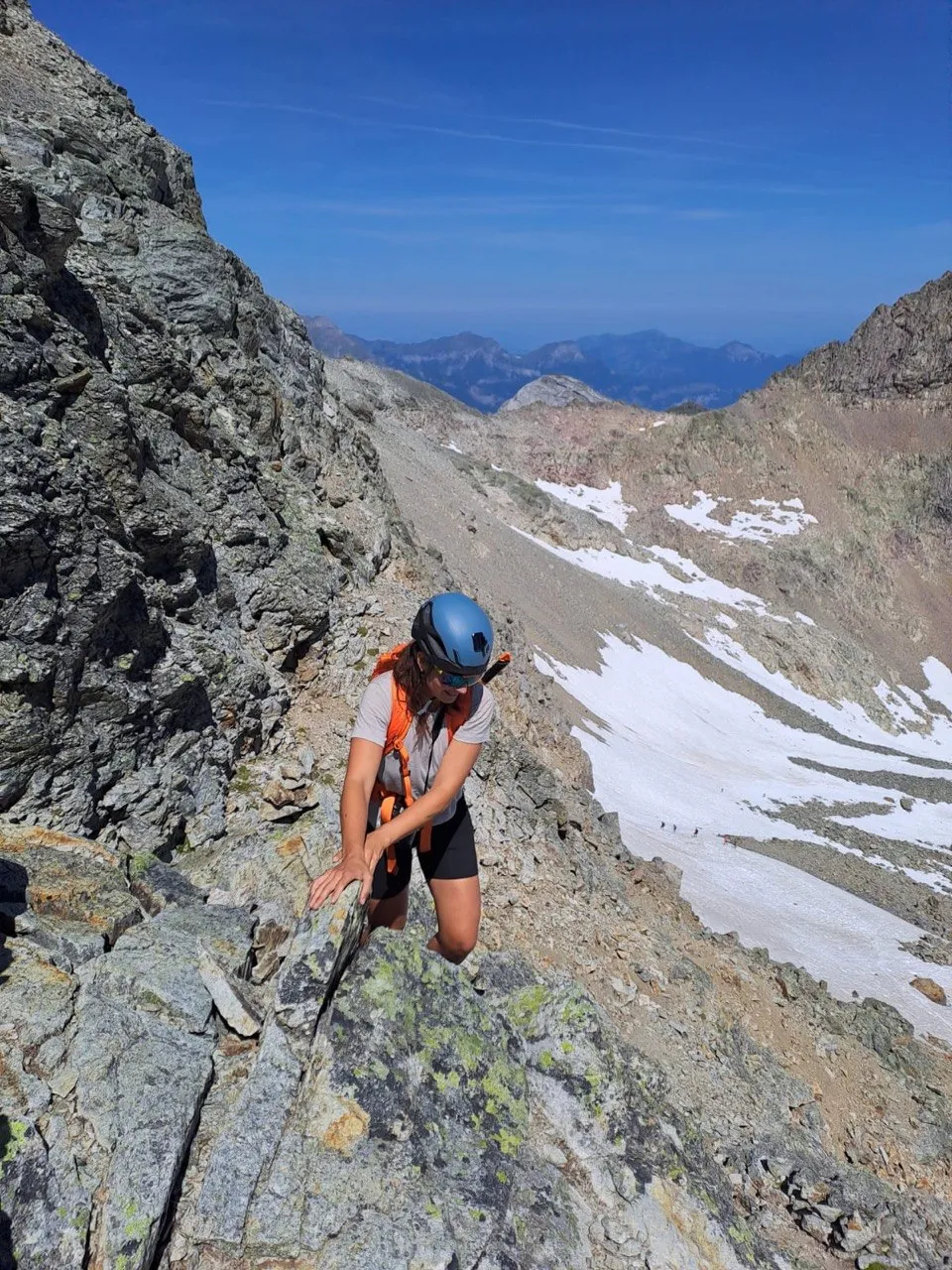 A hiker climbs carefully across an exposed rock passage, a valley with snowfields below.