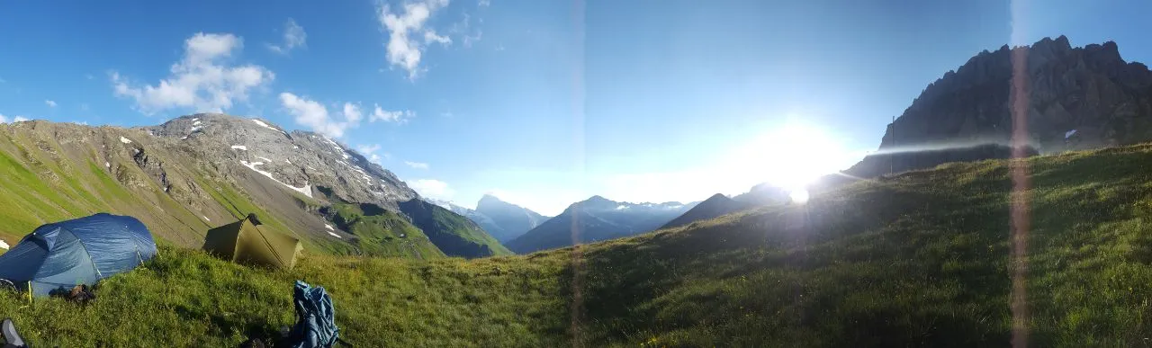 Two tents on an alpine meadow at sunrise, the sun rising beside a jagged rock ridge.