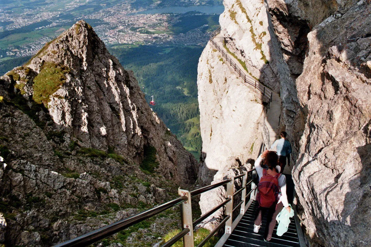 A steep metal staircase cut into the rock near the summit, a red cable car and the city of Lucerne far below.