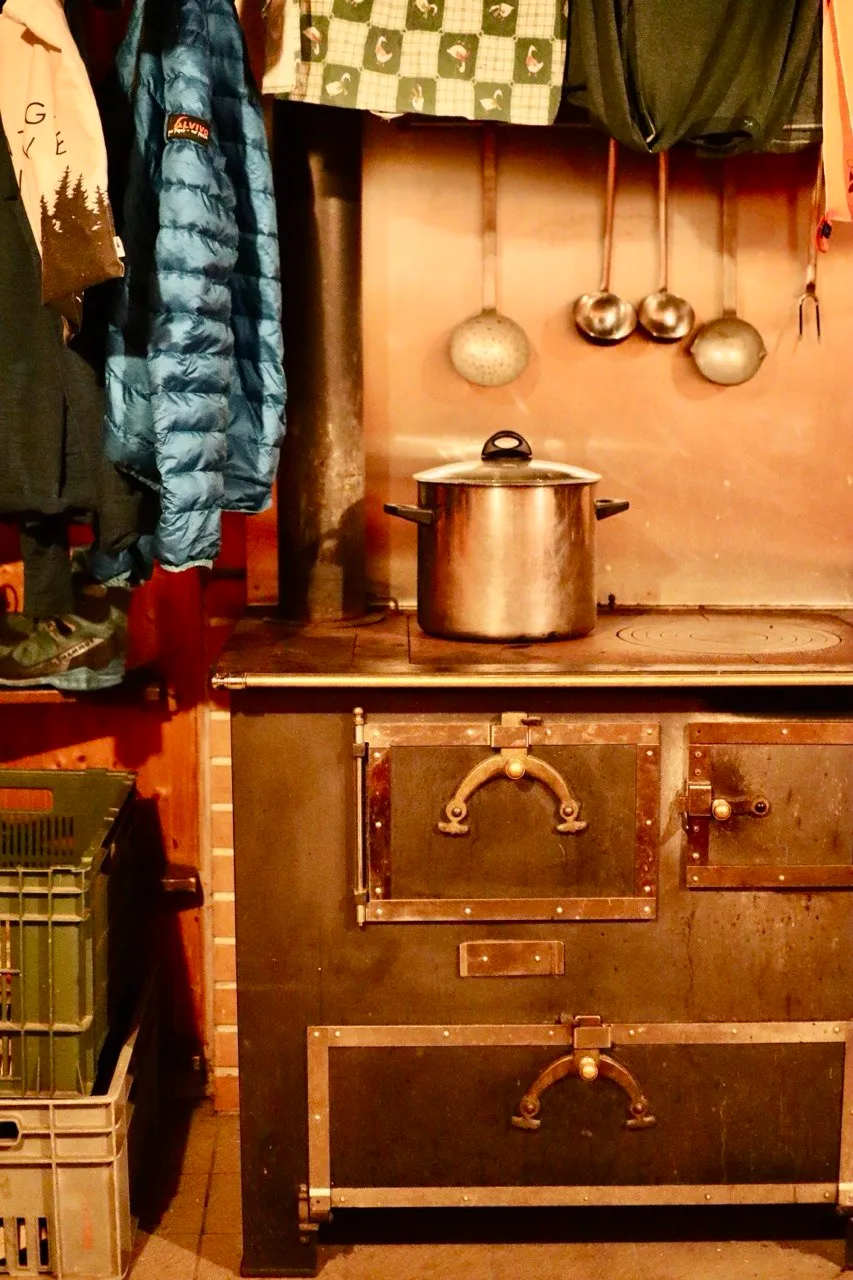 Vintage wood-fired hut stove with a big pot and hanging ladles, jackets drying above.
