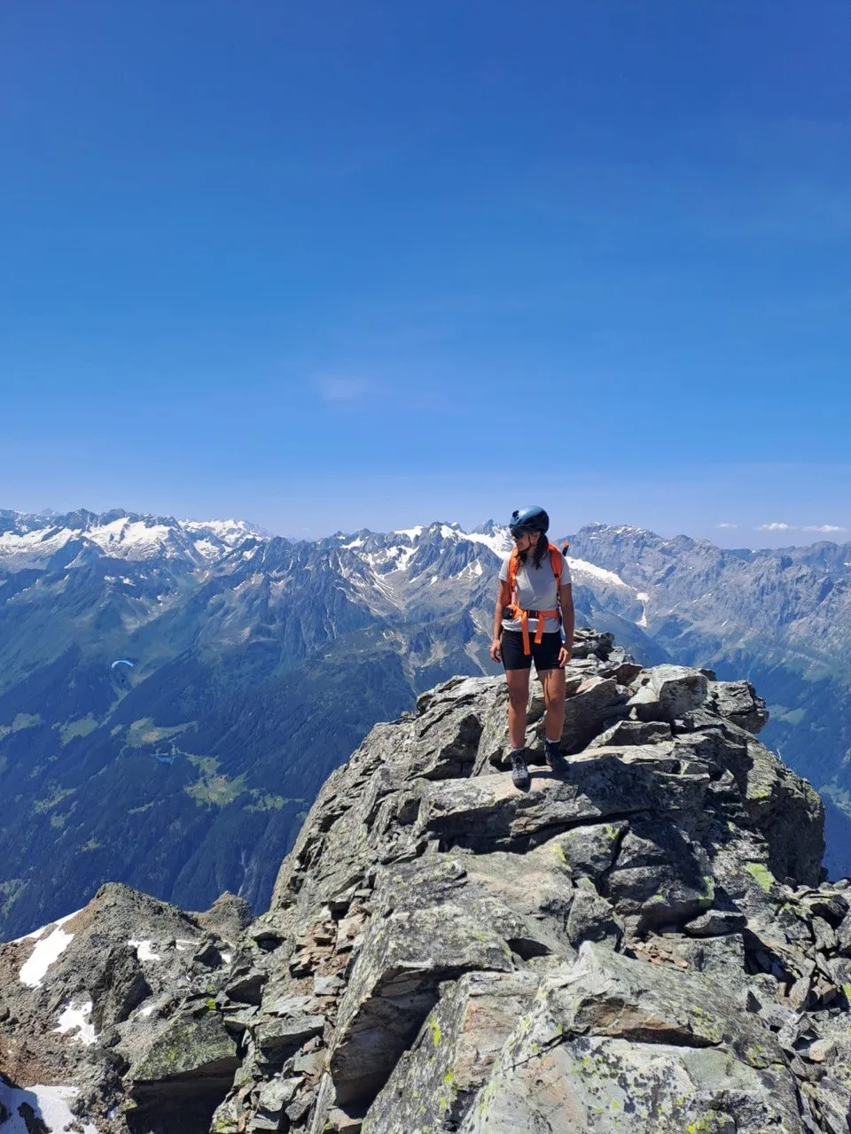 A hiker stands on a narrow rocky summit point, surrounded by a vast panorama of snow-capped Alpine peaks under a deep blue sky.