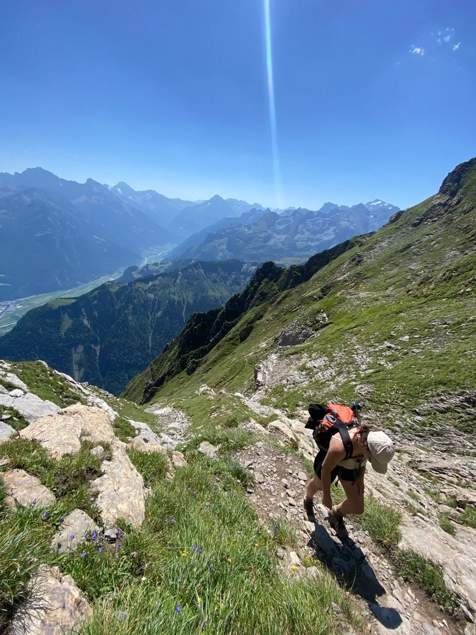 A hiker with a backpack climbs a steep, grassy mountain path, with a valley and mountain ranges behind.