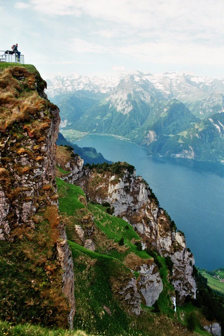 A person leans on the railing of a viewing platform at a grassy cliff edge, the Urnersee and snow-capped peaks across the valley.