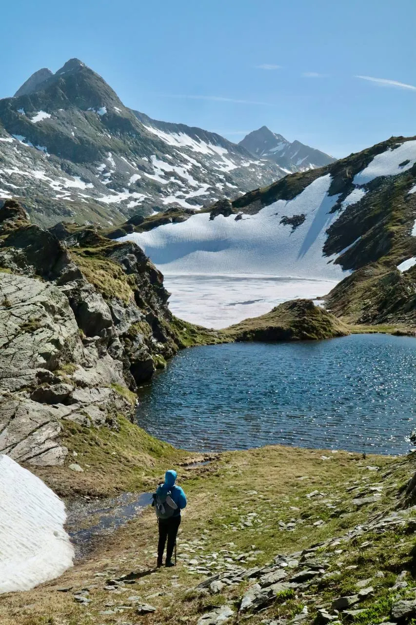 Hiker in a blue jacket on the grassy shore of a small alpine lake below jagged peaks.