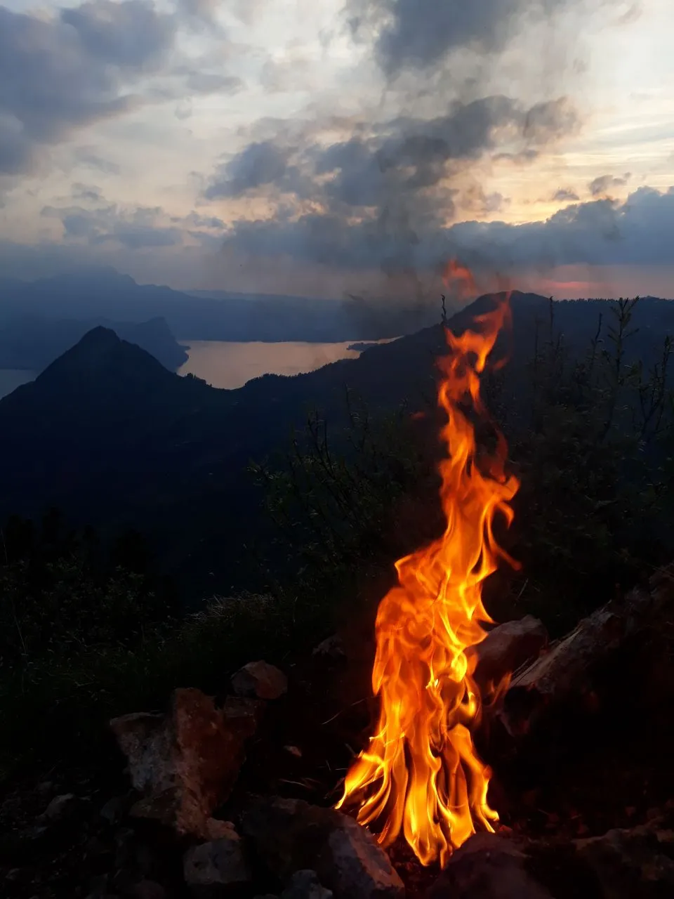 A campfire blazing on the rocky summit at dusk, Lake Lucerne glinting far below under a broken, glowing sky.