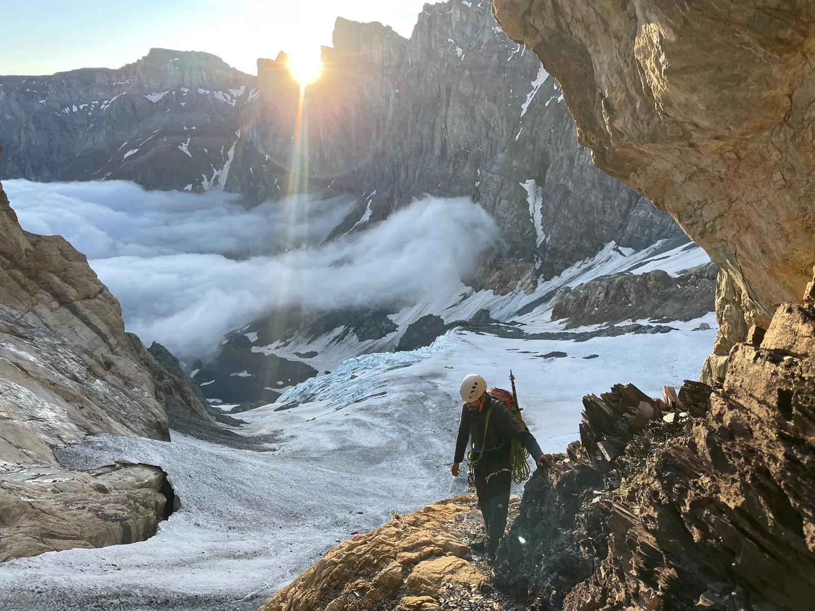 A mountaineer crosses a rocky shoulder backlit by the sun rising over the ridge, with a sea of clouds and the glacier far below.