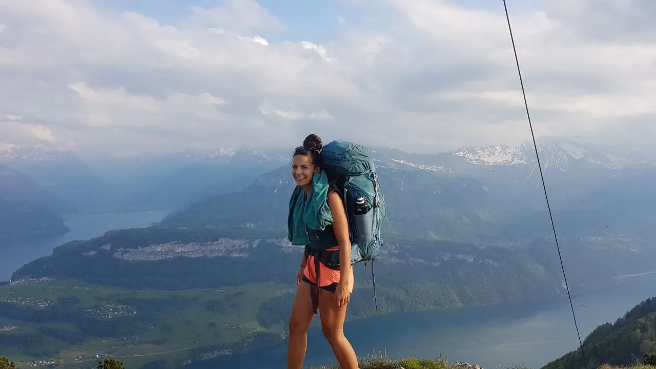 A smiling hiker with a big backpack pauses on the climb, the arms of Lake Lucerne and snow-flecked peaks spread out behind.