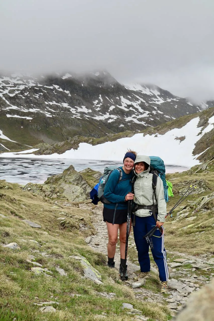 Two hikers with backpacks grinning at the camera in front of a misty, snow-streaked pass.