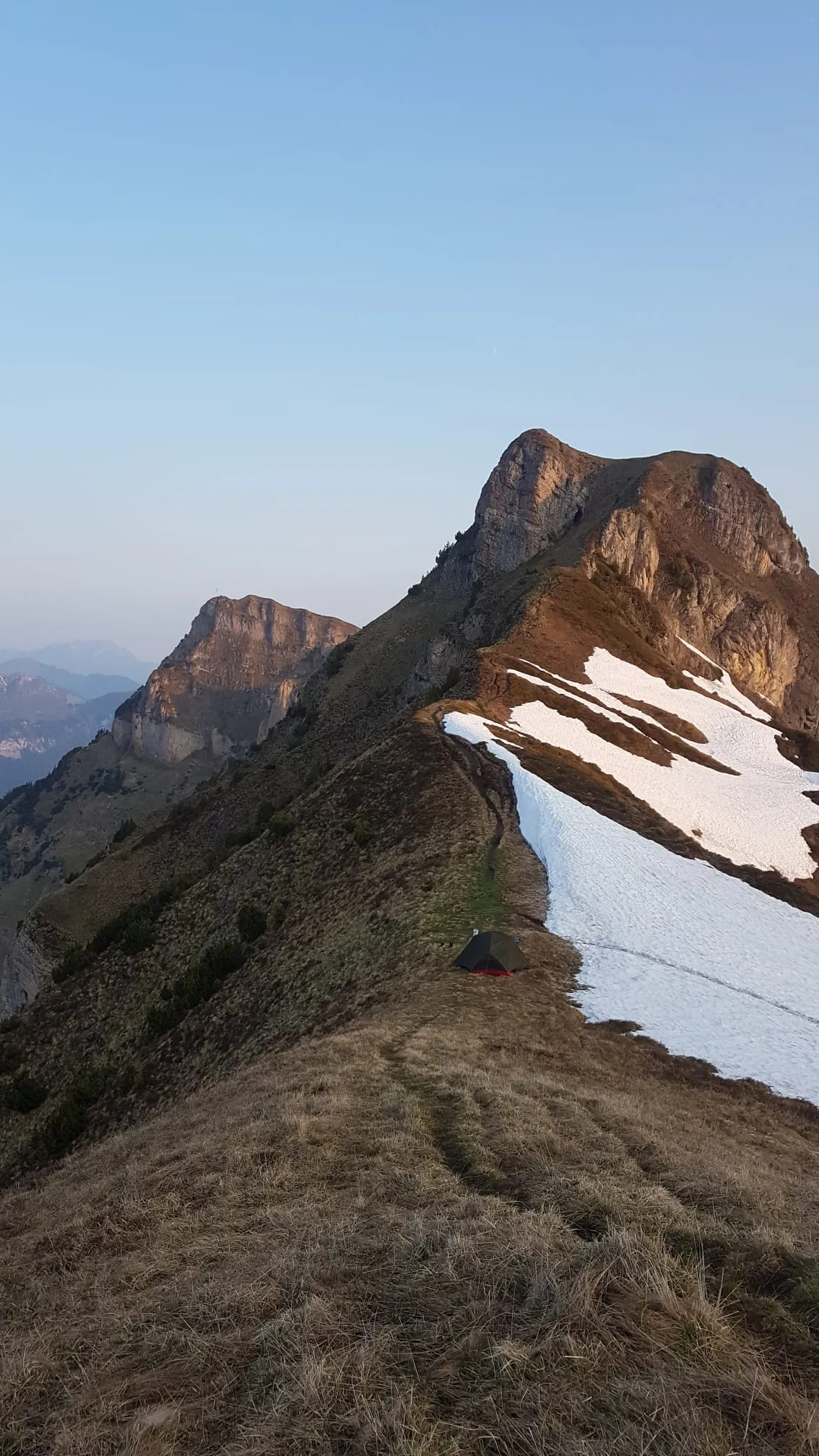 Small tent pitched on a narrow grass ridge beside a snow patch at golden hour.