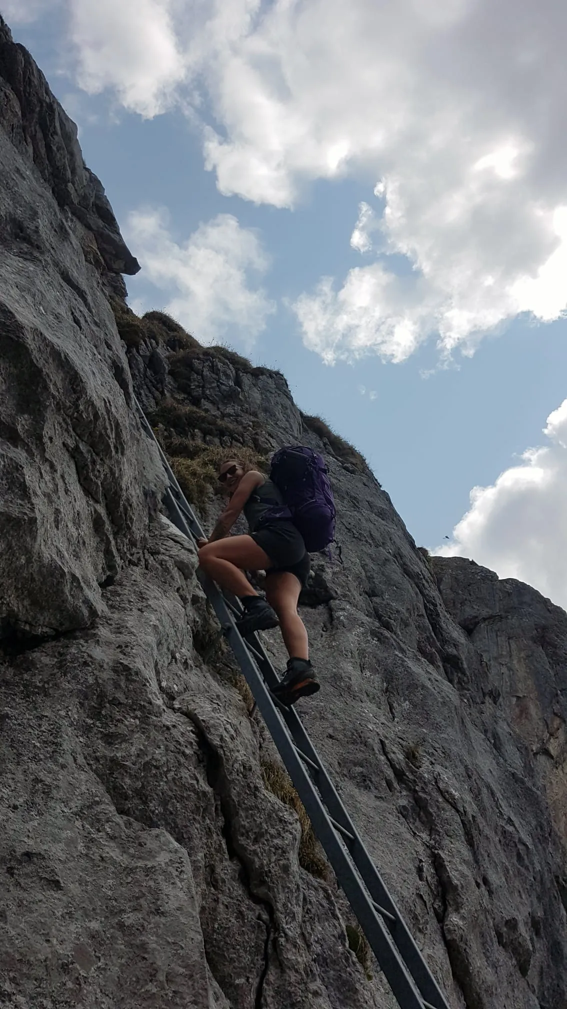 Climbing a metal ladder bolted into a vertical limestone face with a big purple pack.